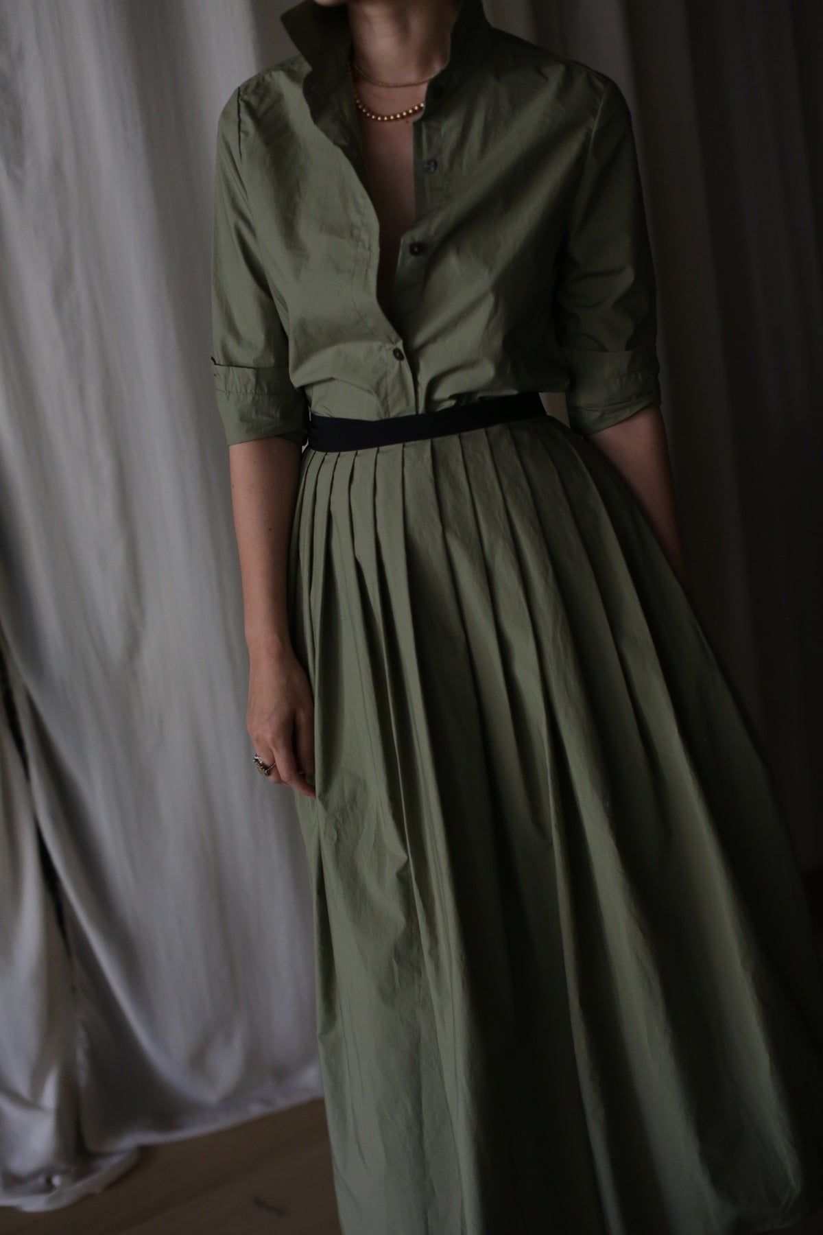 A person wearing the olive Typewriter Wrap Skirt with a black belt and pearl necklace stands before cream-colored curtains. The collared, buttoned long dress features a pleated skirt. Their face is not visible.