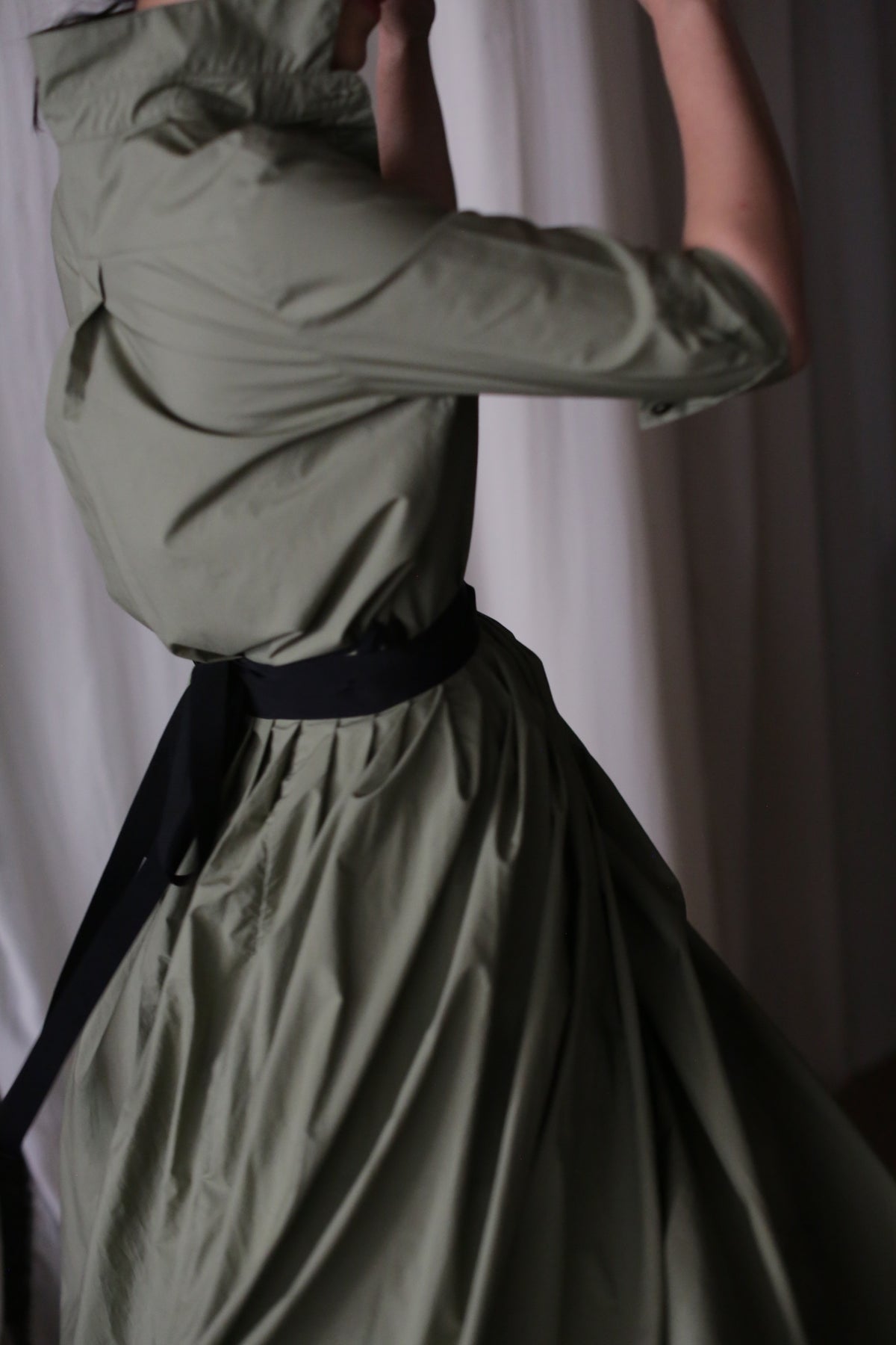 A person wearing an olive green Typewriter Wrap Skirt is captured mid-movement, partially turned away from the camera in soft lighting against a neutral background.