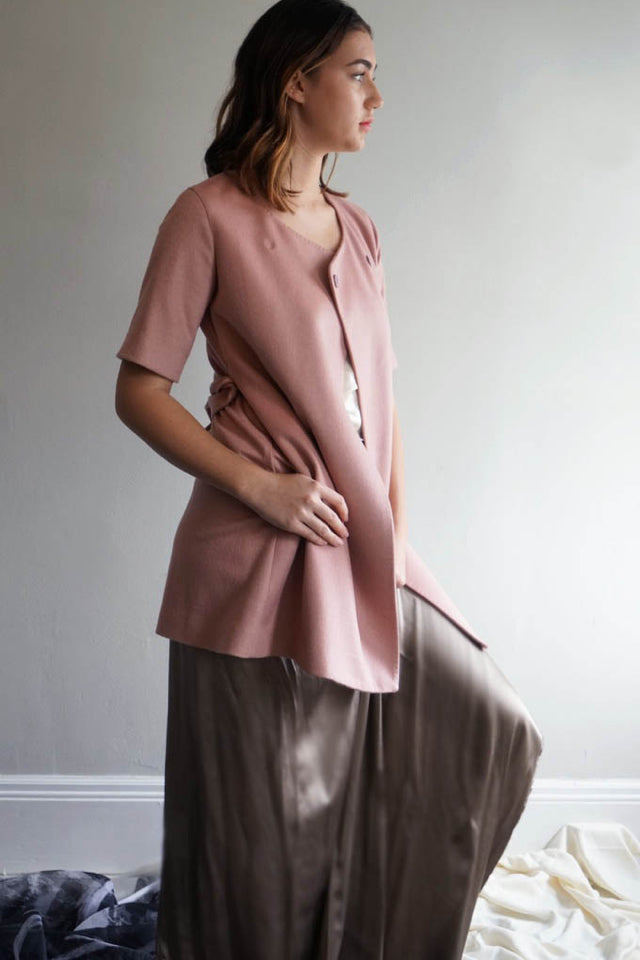 A person wearing the Wexley double-breasted coat over a pink top, paired with wide-legged brown pants, stands indoors. They have shoulder-length brown hair and face sideways, gazing out of view against a light gray wall with a white-framed baseboard.