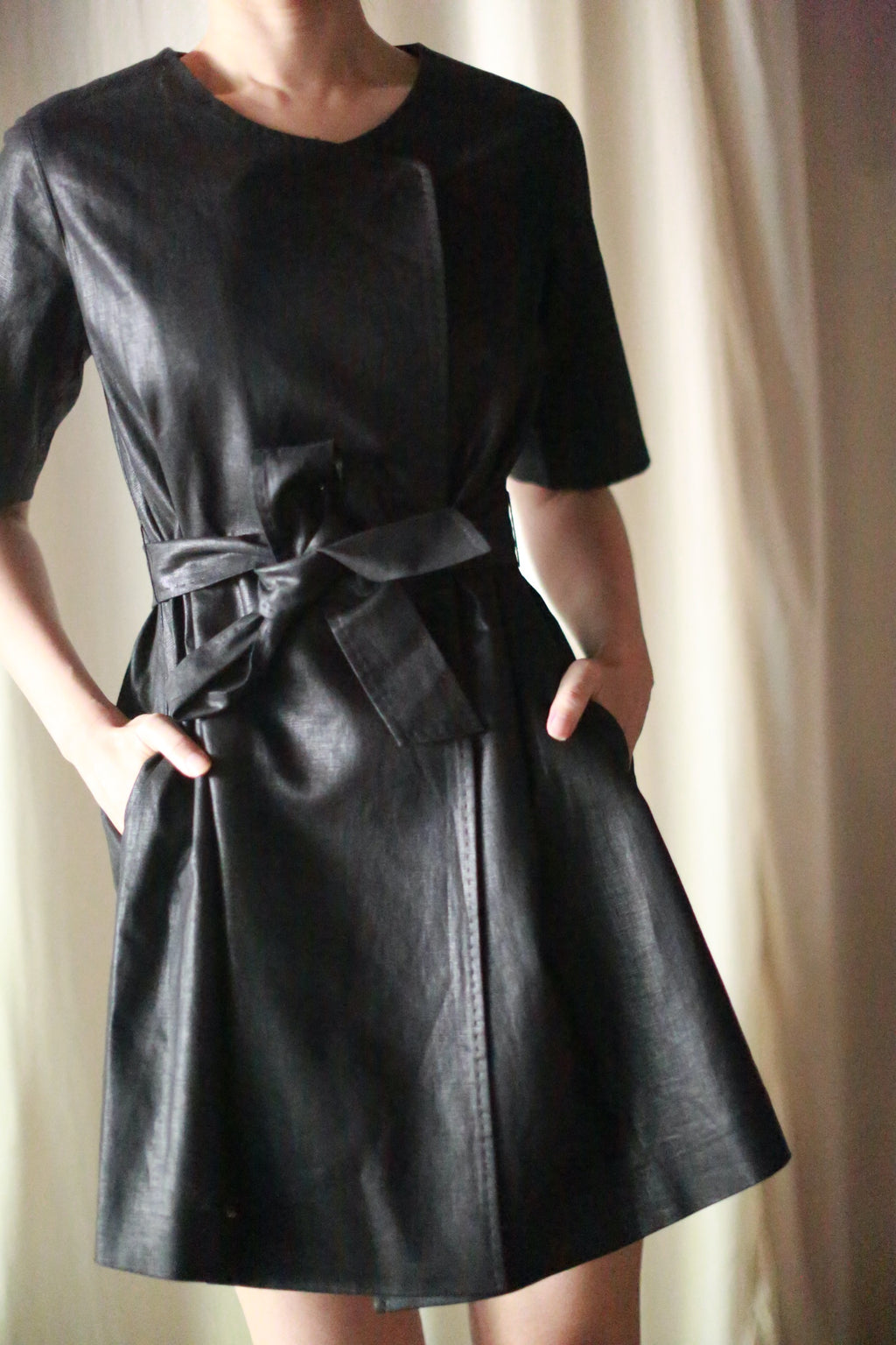 Person wearing the Waxed Linen Wexley, a black short-sleeved leather dress with a tied belt, hands in pockets, standing against a light-colored curtain background; this versatile piece adds an effortlessly chic touch.