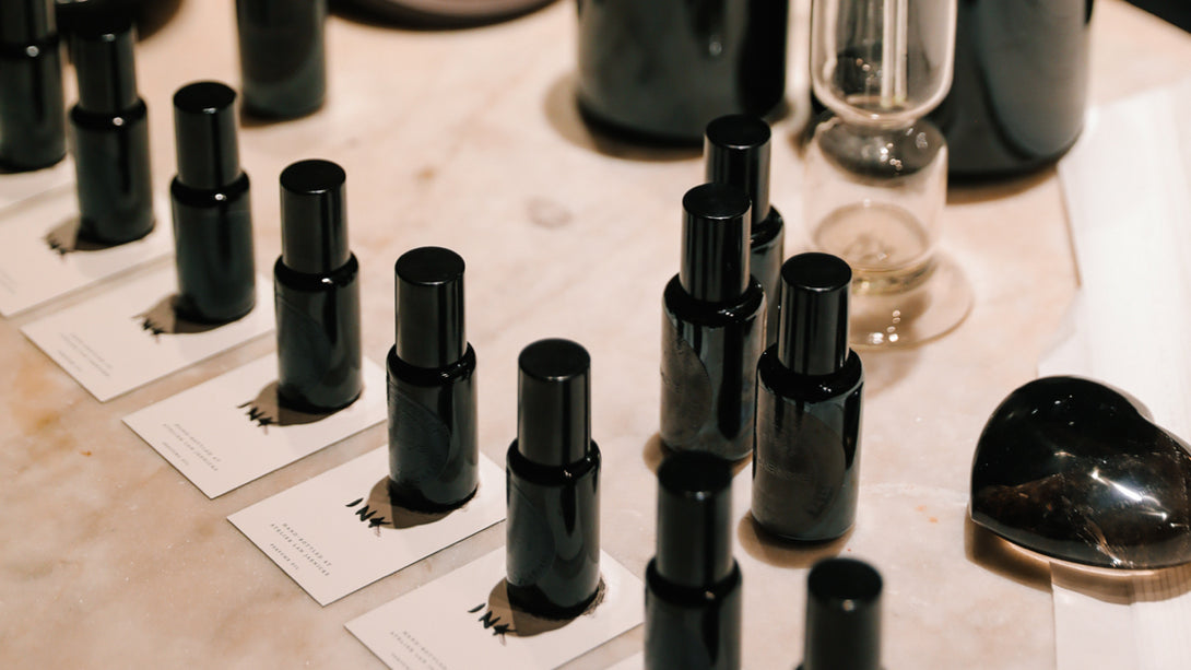 A display of black cosmetic bottles arranged in rows on a marble countertop in a scholar's study. Each bottle sits on a white card with the text 