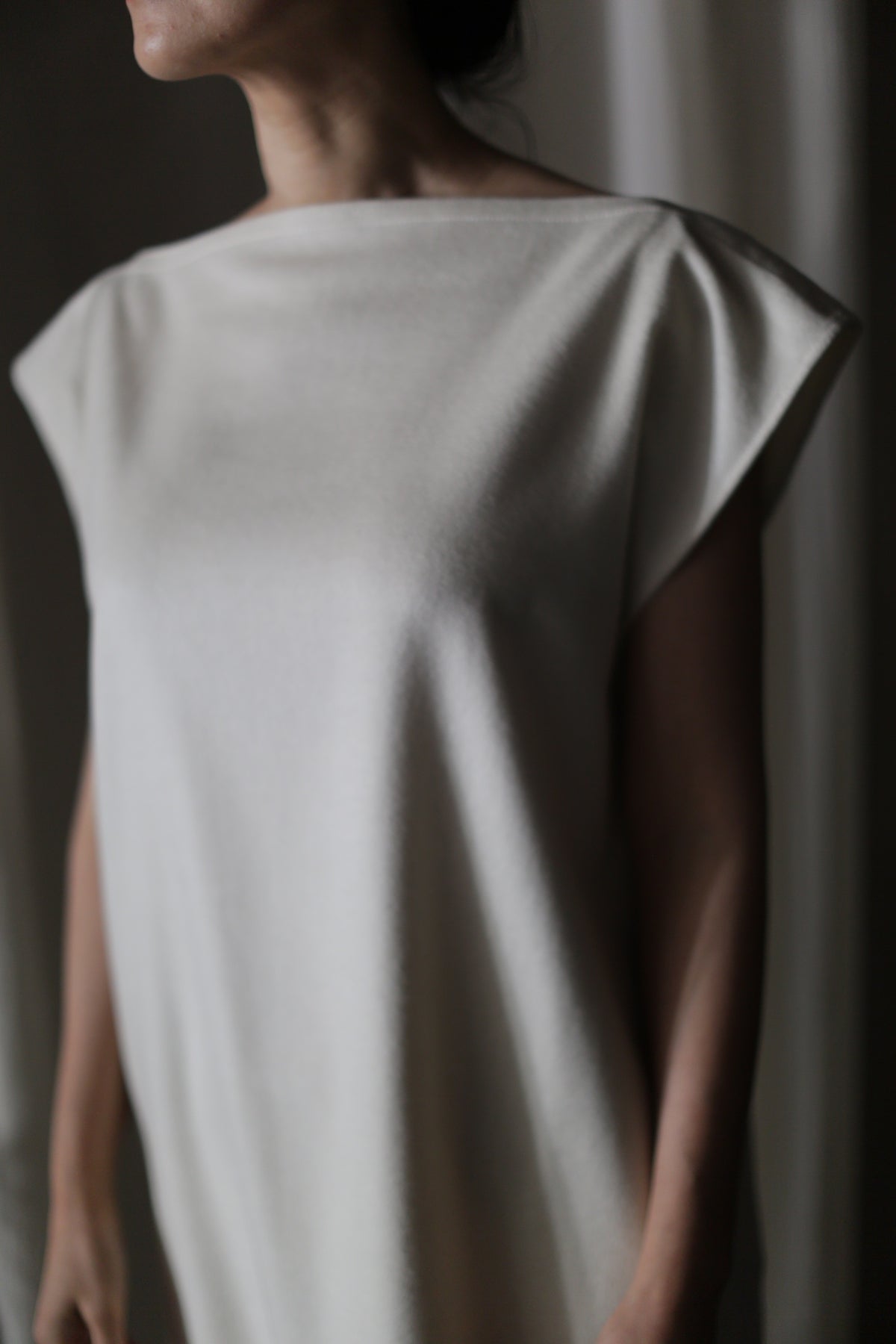 A person is wearing the Paix dress, a simple, sleeveless, light-colored garment adorned with delicate lambswool ruffle detailing. The image captures the upper body and shoulders, with soft lighting creating gentle shadows. The individual's face is not visible. The background is blurred and neutral.