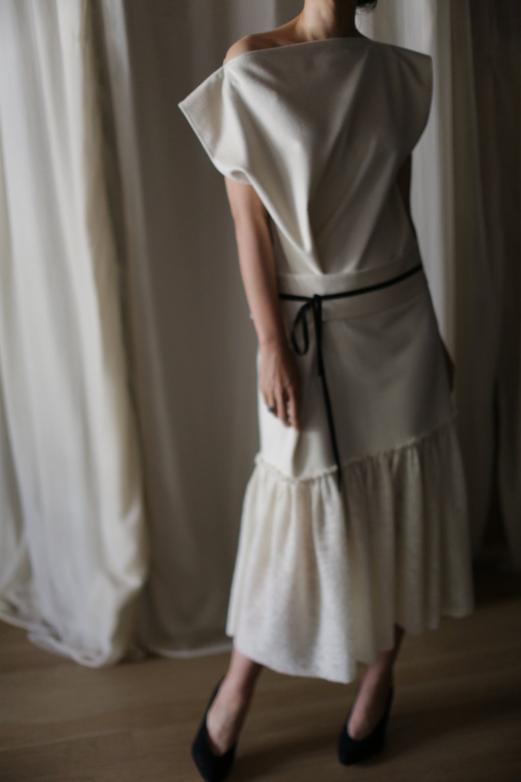 In a softly lit room, a person stands wearing the Paix, an off-the-shoulder white asymmetrical dress with a black belt at the waist. This dress, expertly crafted from woven cashmere, boasts a ruffled hemline. Complementing the outfit are black high-heeled shoes. The background is adorned with light-colored curtains.