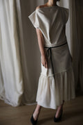 In a softly lit room, a person stands wearing the Paix, an off-the-shoulder white asymmetrical dress with a black belt at the waist. This dress, expertly crafted from woven cashmere, boasts a ruffled hemline. Complementing the outfit are black high-heeled shoes. The background is adorned with light-colored curtains.