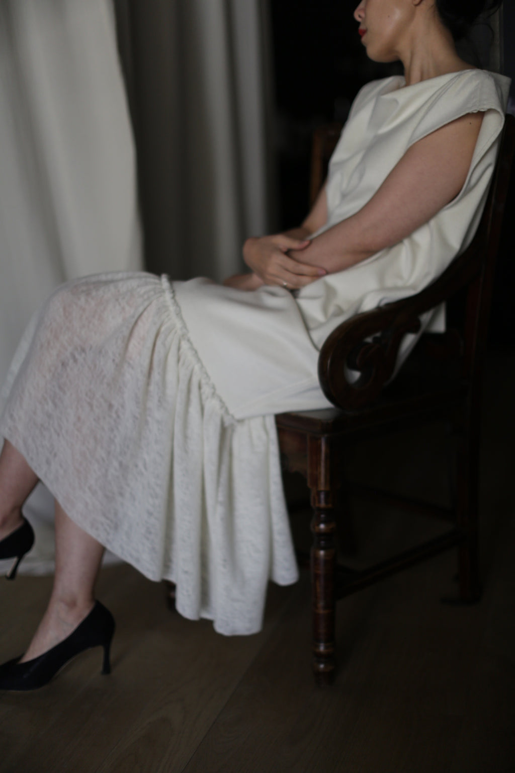 A person is sitting on the Paix wooden chair with their hands resting on their lap. They are wearing a textured, white dress featuring delicate lambswool ruffles and black high heels. The background appears to be neutral and subdued, consisting of fabric drapes. Their face is not fully visible.