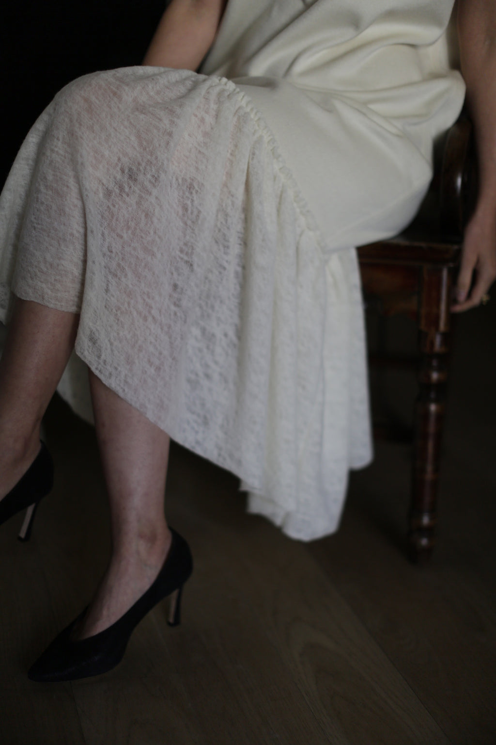 A person wearing the Paix sheer white skirt with a delicate lambswool ruffle and black high heels sits on a wooden chair. Their right hand rests on their lap while their left hand dangles beside them. The person’s legs are slightly crossed, and the lower part of the skirt drapes over the chair.