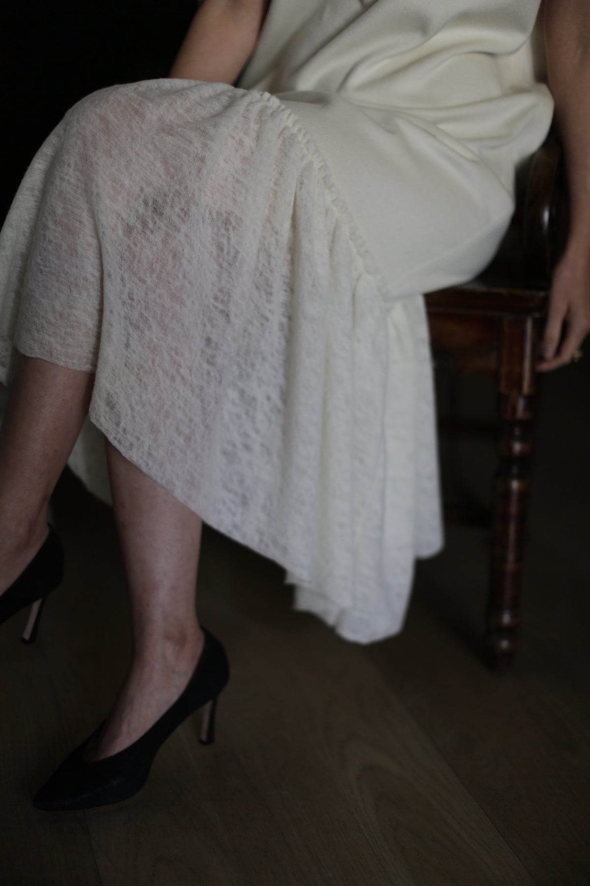 A person wearing the Paix sheer white skirt with a delicate lambswool ruffle and black high heels sits on a wooden chair. Their right hand rests on their lap while their left hand dangles beside them. The person’s legs are slightly crossed, and the lower part of the skirt drapes over the chair.